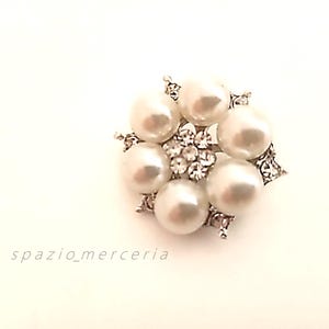 Peut inclure: Broche ronde argentée avec un groupe central de petits cristaux transparents entouré de six grandes fausses perles blanc crème. La broche a un motif floral avec des accents de cristal et le texte "spazio_merceria".