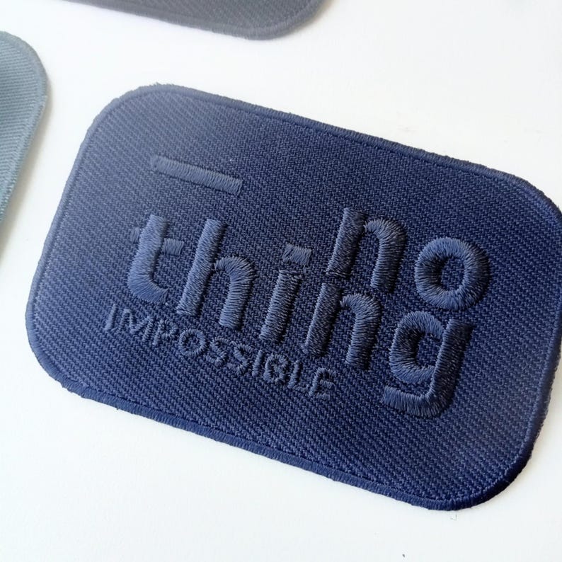 May include: A navy blue rectangular patch with rounded corners. The patch features the embroidered words "nothing impossible" in a raised, textured font. The patch is made of a woven fabric.