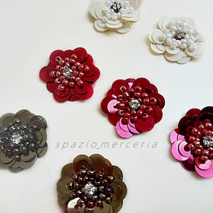 Può includere: Un assortimento di abbellimenti floreali in paillettes di vari colori, tra cui rosso, bianco e marrone. Ogni fiore presenta un cristallo centrale ed è circondato da paillettes. Il testo "spazio_merceria" è visibile.