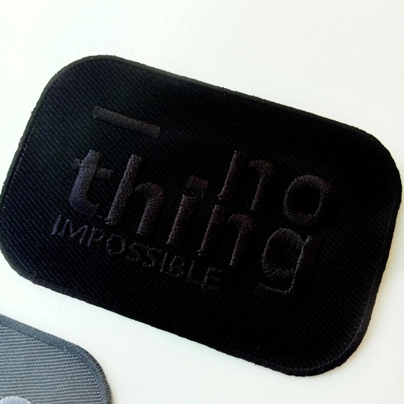 May include: Black rectangular patch with the embroidered text "thing impossible" in a dark tone. The patch has a textured surface and a stitched border. The patch is on a white background.