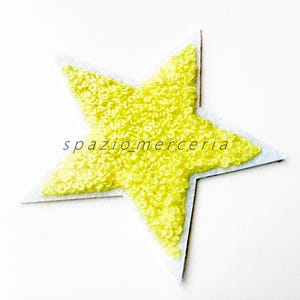 Può includere: Toppa di stoffa a forma di stella gialla con superficie strutturata e ad anello. La stella è delineata da un bordo grigio chiaro e presenta il testo "spazio merceria" in stile scritto a mano.