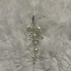 May include: A silver-toned phone charm featuring a white bow, two white faux pearls, a pair of silver angel wings, a silver heart frame, and a small silver key charm with wings at the top, all connected by a thin white cord loop.