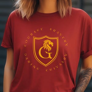 May include: Cardinal red t-shirt with a gold crest design featuring a lion and the letter "G". The crest is surrounded by the words "Courage, Bravery, Daring, Chivalry". The word "CARDINAL" is printed at the bottom.