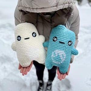 May include: Two handmade crochet plush toys. One is off-white, the other is light blue. Both have black eyes and a stitched mouth. The blue toy has a white design on its belly. The toys are held in front of a snowy background.