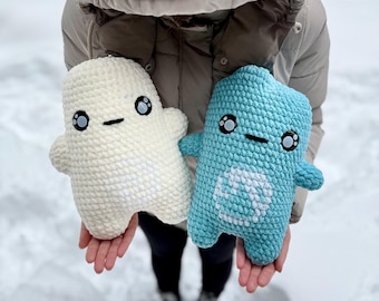Può includere: Due peluche fatti a mano all'uncinetto. Uno è bianco sporco, l'altro è azzurro. Entrambi hanno occhi neri e una bocca cucita. Il giocattolo blu ha un disegno bianco sulla pancia. I giocattoli sono tenuti davanti a uno sfondo innevato.
