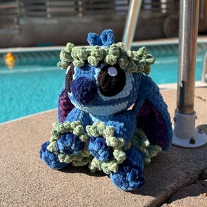 May include: A handmade blue and green crochet plush toy, featuring large black eyes, a blue nose, and a green leafy crown. The plush is sitting on a concrete surface near a pool.