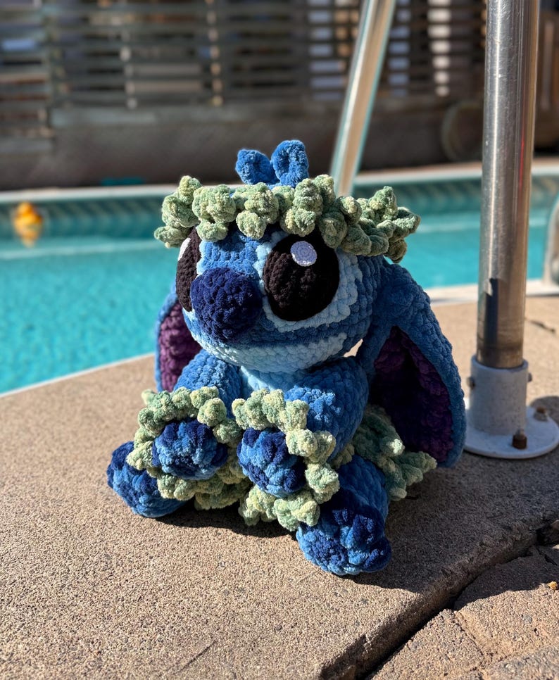 May include: A handmade blue and green crochet plush toy, featuring large black eyes, a blue nose, and a green leafy crown. The plush is sitting on a concrete surface near a pool.