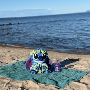 May include: A blue and purple crochet Stitch doll with a green floral crown sits on a teal towel on a sandy beach. A purple cup with a flamingo design and a straw is next to the doll. The ocean and sky are in the background.