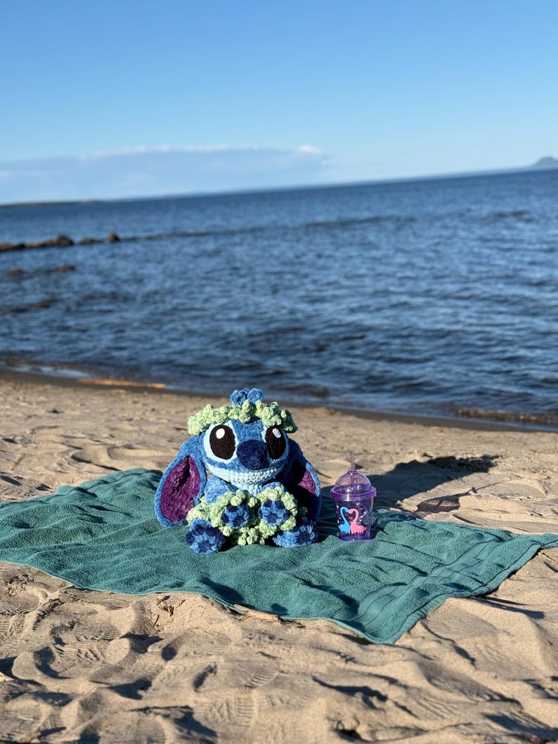 May include: A blue and purple crochet Stitch doll with a green floral crown sits on a teal towel on a sandy beach. A purple cup with a flamingo design and a straw is next to the doll. The ocean and sky are in the background.