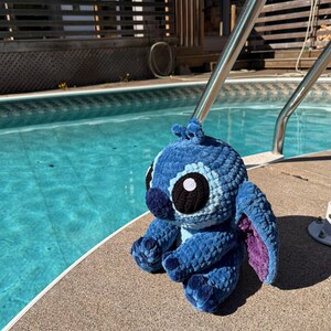 May include: A blue crochet plush toy of a cartoon character with large black and white eyes, and large ears with purple insides. The plush is sitting on the edge of a pool.