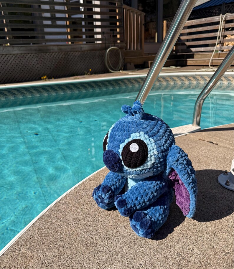 May include: A blue crochet plush toy of a cartoon character with large black and white eyes, and large ears with purple insides. The plush is sitting on the edge of a pool.