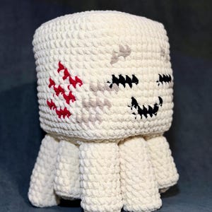 Puede incluir: Un juguete de peluche de ganchillo hecho a mano, de color blanquecino, con una cabeza cuadrada y cuatro patas cortas. La cara presenta ojos y boca cosidos en negro, con acentos rojos y grises. El juguete tiene un aspecto texturizado y suave.