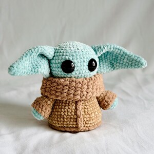 May include: A crocheted plush toy resembling a character with large black eyes and large, pointed ears. The head and ears are light blue, the body is brown, and the coat is a lighter brown. The toy is approximately 15 cm tall.