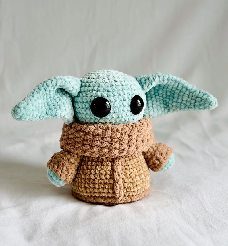May include: A crocheted plush toy resembling a character with large black eyes and large, pointed ears. The head and ears are light blue, the body is brown, and the coat is a lighter brown. The toy is approximately 15 cm tall.