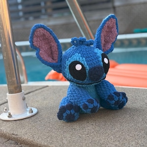 May include: A handmade blue crochet Stitch doll with large ears, black eyes, and a small nose. The plush toy is sitting on a concrete surface near a pool. The ears have a pink interior.