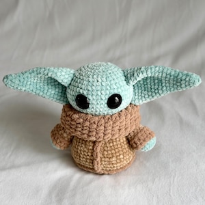 May include: A crocheted plush toy of a character with large black eyes, large ears, and a tan robe. The toy's head and ears are light blue, and the robe is tan. The toy is sitting on a white surface.
