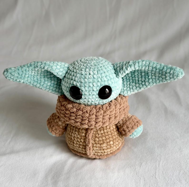 May include: A crocheted plush toy of a character with large black eyes, large ears, and a tan robe. The toy's head and ears are light blue, and the robe is tan. The toy is sitting on a white surface.
