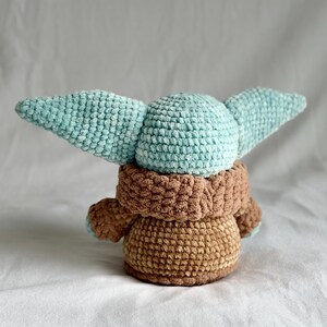 May include: A crocheted doll with a light blue head and large ears, a brown robe, and a tan body. The soft toy is made with textured yarn, giving it a cuddly appearance. The doll is a character from a popular science fiction franchise.