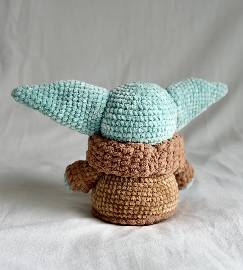 May include: A crocheted doll with a light blue head and large ears, a brown robe, and a tan body. The soft toy is made with textured yarn, giving it a cuddly appearance. The doll is a character from a popular science fiction franchise.