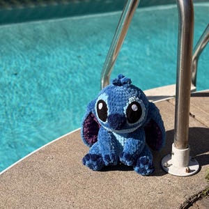 May include: A blue crochet plush toy with large black eyes, white highlights, and purple inner ears. The plush is sitting on a concrete surface next to a pool with a stainless steel handrail.