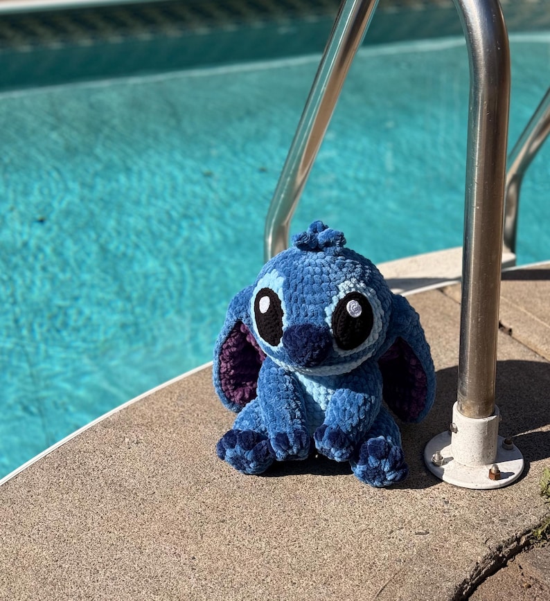 May include: A blue crochet plush toy with large black eyes, white highlights, and purple inner ears. The plush is sitting on a concrete surface next to a pool with a stainless steel handrail.