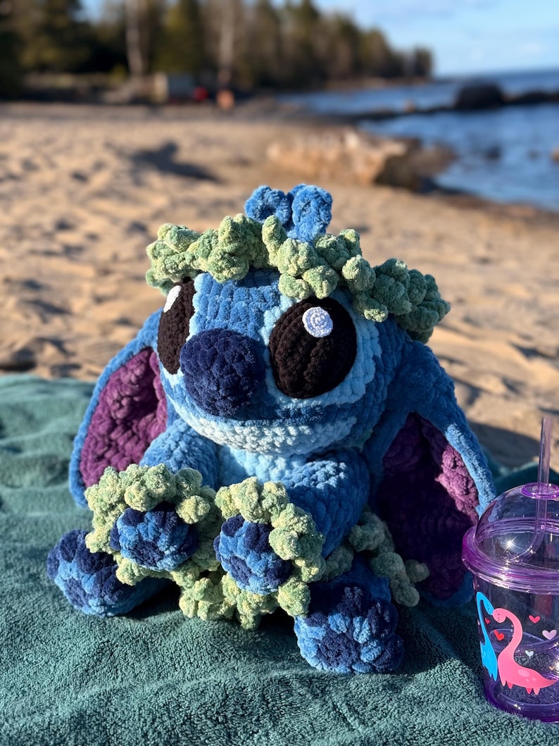 May include: A handmade blue crochet plush toy with large black eyes, a navy blue nose, and green accents. The plush is sitting on a teal blanket near the beach. A purple cup with a dinosaur design is also visible.