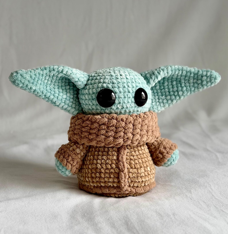 May include: A handmade crochet doll resembling a popular character. The doll has large black eyes and oversized ears, in teal and brown. The body is brown, with a teal head and ears, and a brown collar. A soft toy.