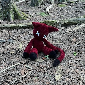 May include: A handmade, dark red crochet cat doll with black accents. The cat has white cross-shaped eyes and is sitting on the forest floor. The tail and feet are black. The doll has pointed ears and a long tail.