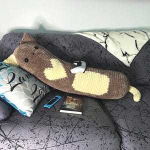 Może przedstawiać: Poduszka w kształcie kota wykonana na szydełku w kolorze brązowym i żółtym z sercowatymi detalami leży na szarej kanapie. Na kanapie leży również biały kontroler do gier wideo i książka.