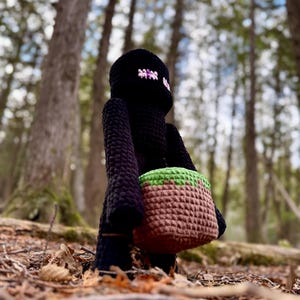 May include: A black crocheted Minecraft Creeper plush toy holding a brown and green crocheted dirt block. The toy is standing in a woodland setting.