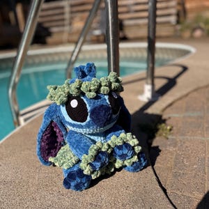 May include: A handmade blue crochet plush toy with large black eyes, a blue nose, and a green floral crown. The plush has large ears with purple lining and green accents. It is sitting on a concrete surface near a pool.