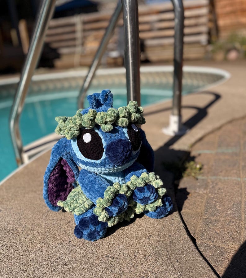 May include: A handmade blue crochet plush toy with large black eyes, a blue nose, and a green floral crown. The plush has large ears with purple lining and green accents. It is sitting on a concrete surface near a pool.