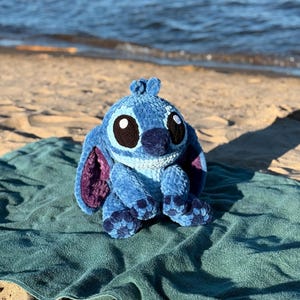 May include: A blue and purple crochet plush toy of a cartoon character with large black eyes, sitting on a teal towel on a sandy beach. The plush has large ears and a textured appearance, with a dark blue nose.