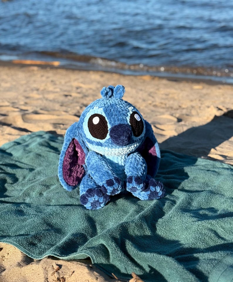 May include: A blue and purple crochet plush toy of a cartoon character with large black eyes, sitting on a teal towel on a sandy beach. The plush has large ears and a textured appearance, with a dark blue nose.