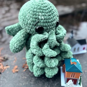 May include: A handmade, mint green crochet octopus plush toy with large black eyes and curled tentacles. The plush is sitting on a gray surface with miniature houses and trees in the background.