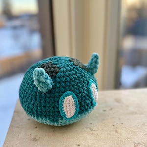 Puede incluir: Un juguete de peluche de color azul turquesa tejido a crochet con detalles en blanco y negro. El juguete tiene dos ojos grandes y dos pequeños cuernos.