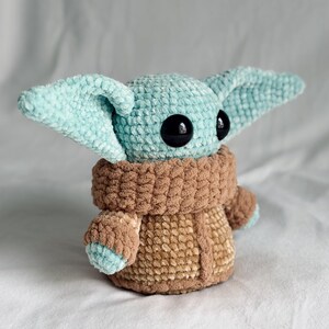 May include: A crocheted plush toy resembling a character with large ears and black eyes. The toy is mainly light blue and brown, with a textured, fuzzy appearance. The character is wearing a brown coat.