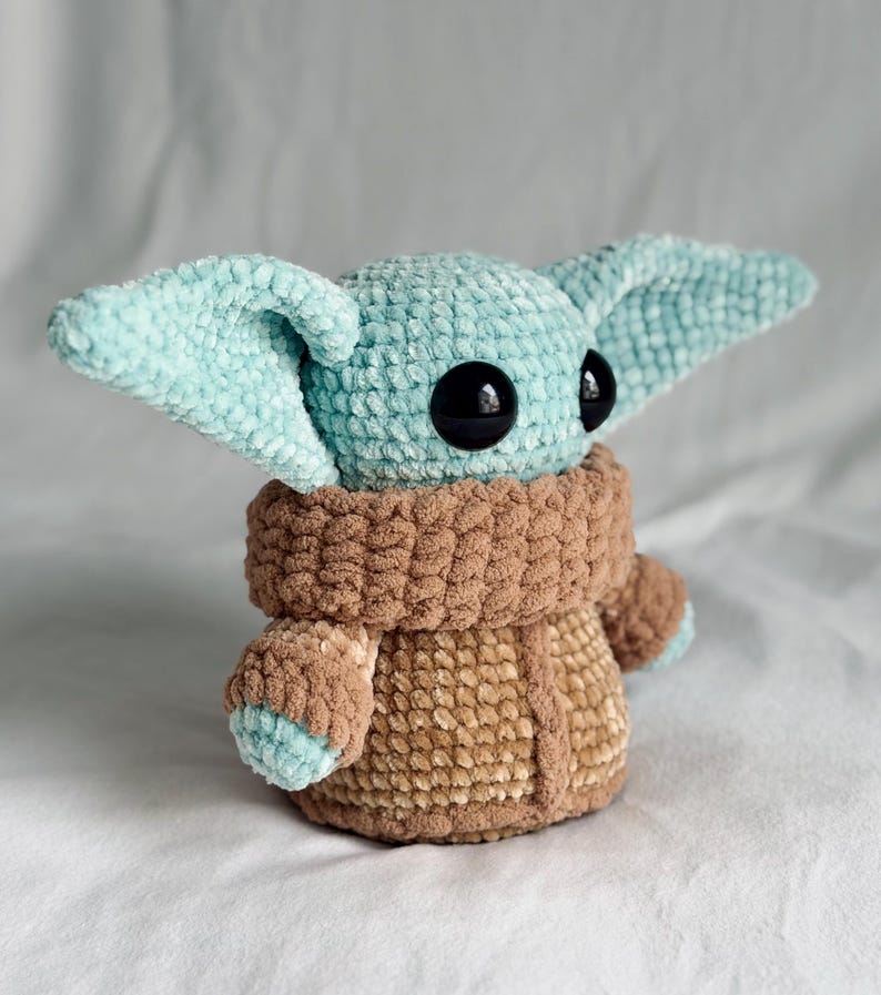 May include: A crocheted plush toy resembling a character with large ears and black eyes. The toy is mainly light blue and brown, with a textured, fuzzy appearance. The character is wearing a brown coat.