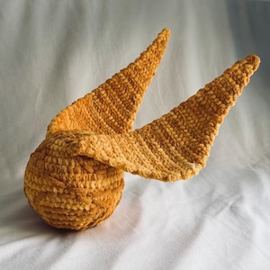 May include: A golden yellow crocheted sphere with two large, textured, wing-like extensions protruding from the sides. The object is crafted from a soft, plush yarn material, featuring a consistent, dense stitch pattern throughout the entire structure.