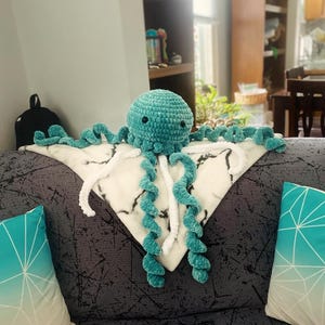 Peut inclure: Une peluche de méduse turquoise crochetée avec de longs tentacules bouclés, posée sur une couverture à motifs noirs et blancs. La peluche est sur un canapé gris avec deux coussins à motifs géométriques turquoise et blancs.