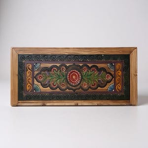 May include: A rectangular wooden box with a hand-painted floral design. The central motif features a red flower surrounded by green leaves and smaller red flowers. The border is dark green and purple, with a natural wood frame.