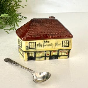 May include: A ceramic miniature of "The Old Curiosity Shop" with a brown roof and yellow walls. A small silver spoon rests in front of the shop. Green foliage is in the background.