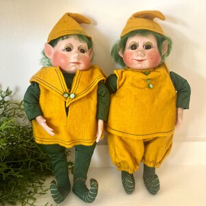 May include: Two vintage elf dolls. Each doll wears a mustard yellow felt outfit with a matching hat and green accents. The dolls have green hair, rosy cheeks, and are posed side-by-side.