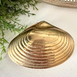 May include: A golden, clam-shaped decorative dish. The dish has a ridged texture and a shiny, reflective surface. The background includes a painting in a decorative frame and some green foliage.