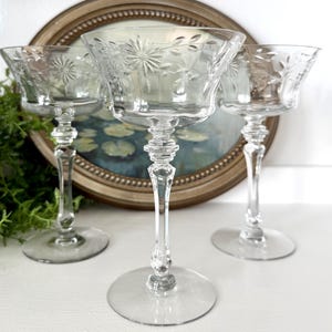 May include: Three clear, stemmed champagne glasses with etched floral designs. The glasses have a wide bowl, a slender stem, and a round base. The background includes a framed painting and greenery, creating a vintage aesthetic.