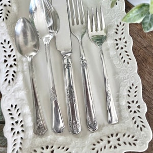 May include: A set of five silver-plated utensils, including a butter knife, two forks, and two spoons, arranged on a white lace doily.