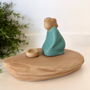 May include: A ceramic sculpture featuring a figure in a teal-colored cloak, seated on a light brown, organic-shaped base. A small, round, beige object sits beside the figure. The sculpture is set against a white background with greenery.