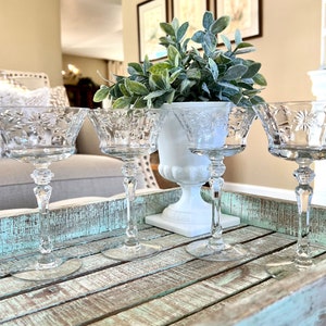 May include: Four clear crystal champagne glasses with starburst designs sit on a distressed, turquoise-painted wooden tray. A white vase with greenery is in the background. The glasses have long stems and detailed bowls.