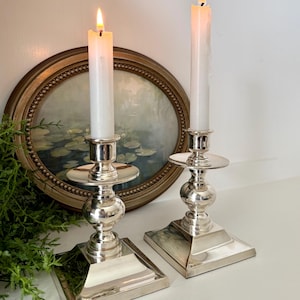 May include: Two silver candle holders with lit white candles. The candle holders have a square base, a round center, and a circular top. An oval framed painting is in the background.