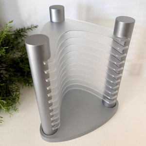 May include: A silver-colored media storage rack featuring three cylindrical vertical supports connected by a triangular base and top. The unit includes multiple clear, curved plastic dividers designed to hold individual discs in an upright position.